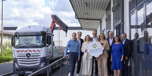Acht Frauen und Männer in formeller Kleidung stehen neben einem silbernen LKW. Zwei von Ihnen halten ein Schild mit der Aufschrift "Innovationspreis Münsterland".