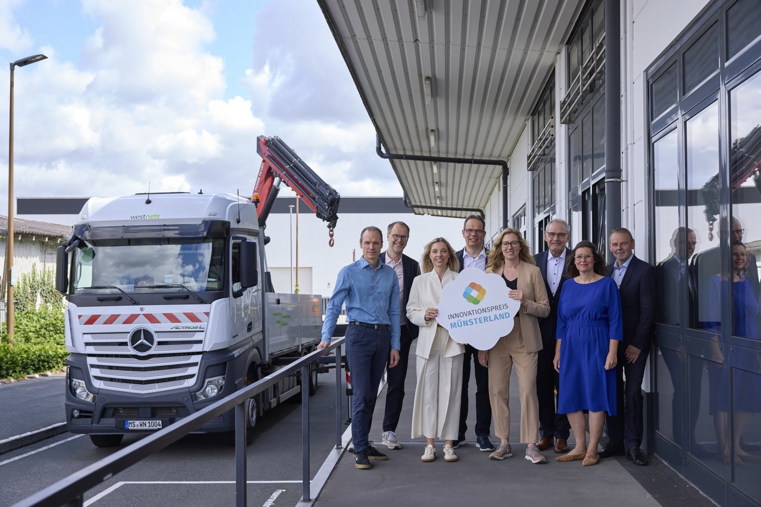 Acht Frauen und Männer in formeller Kleidung stehen neben einem silbernen LKW. Zwei von Ihnen halten ein Schild mit der Aufschrift "Innovationspreis Münsterland".
