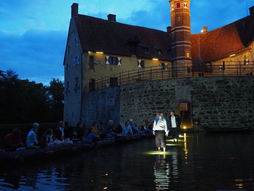 Vor einer Burg laufen zwei historisch gekleidete Personen durch das Wasser des Burggrabens. Links sitzen Zuschauer in kleinen Booten.