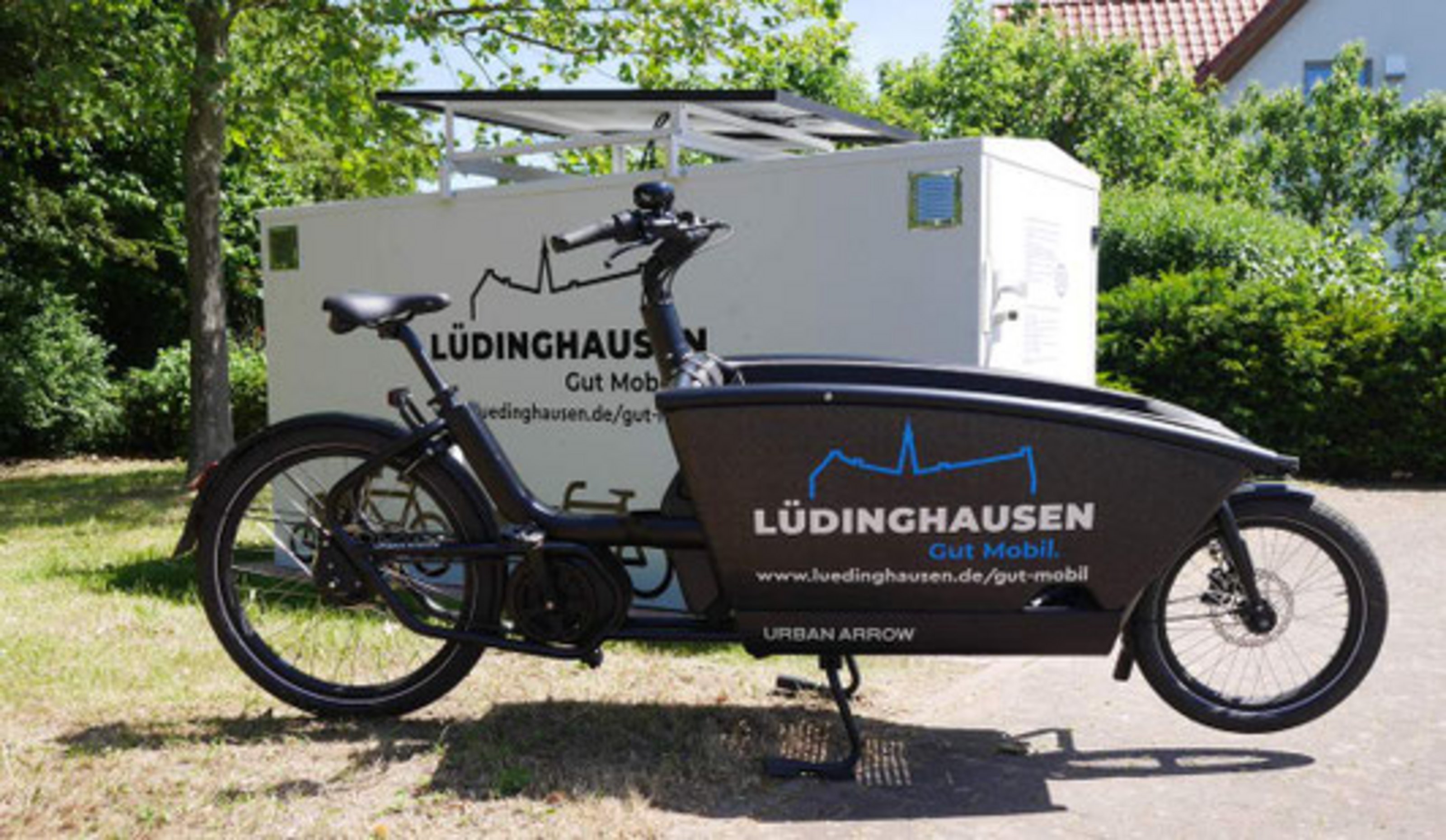 Lastenfahrrad mit Aufbau-Anhänger, beschriftet mit ‚Lüdinghausen – Gut mobil‘, steht im Freien vor Bäumen