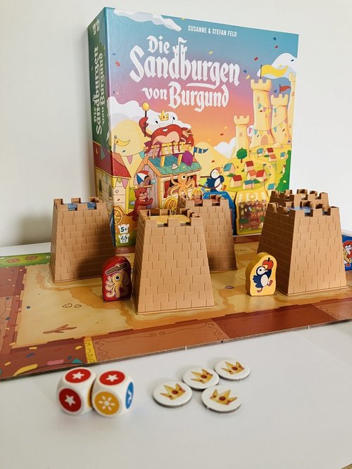 Auf einem Tisch ist ein Brettspiel mit Burgen, Spielsteienen und Würfeln aufgebaut. Dahinter steht ein Spielkarton, auf dem "Die Sandburgen von Burgund" steht.