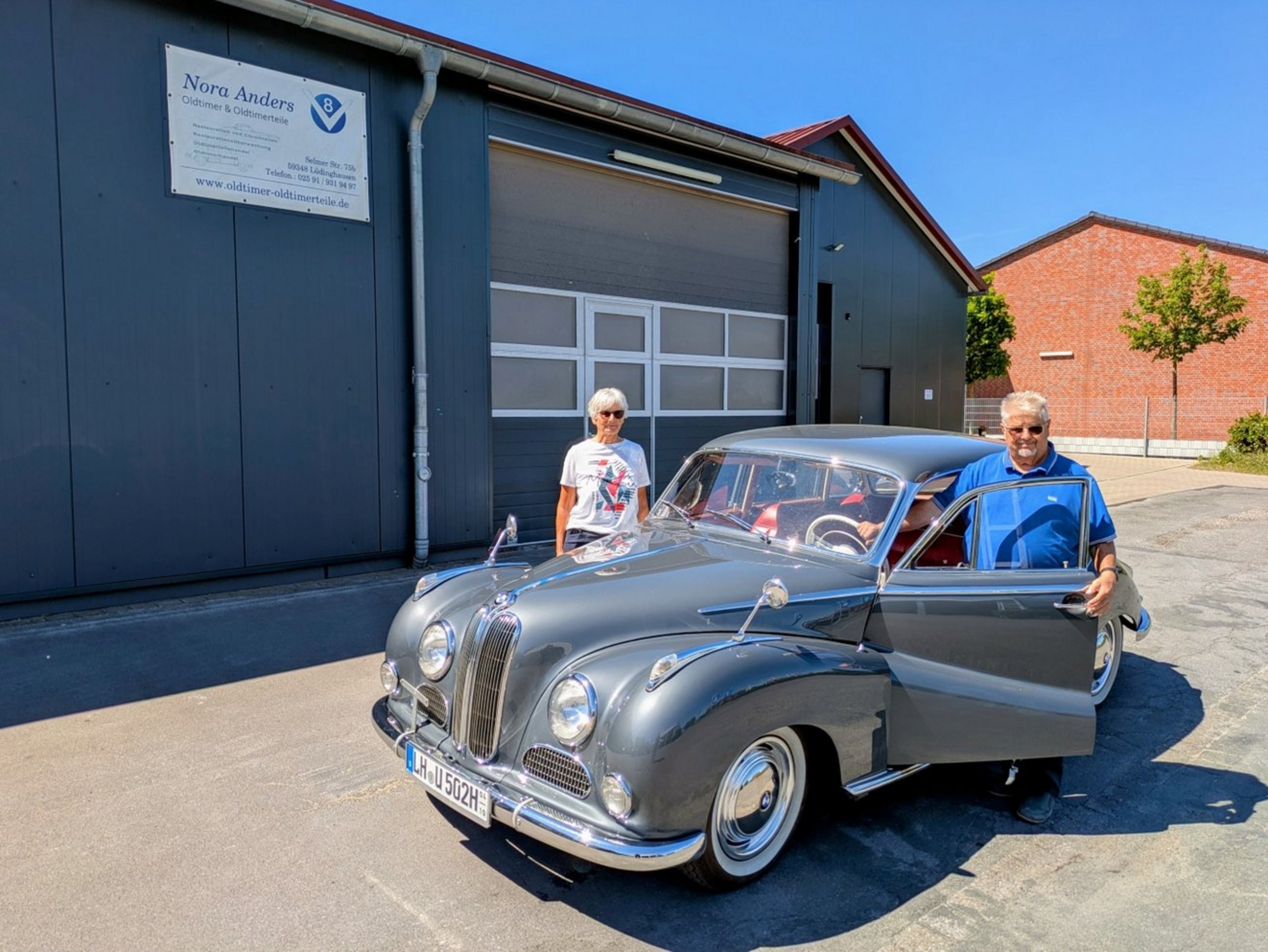 Ursula und Peter Gerding stehen vor einer Werkstatt neben ihrem grauen BMW 502 „Barockengel“, ein Oldtimer mit geöffneter Tür.