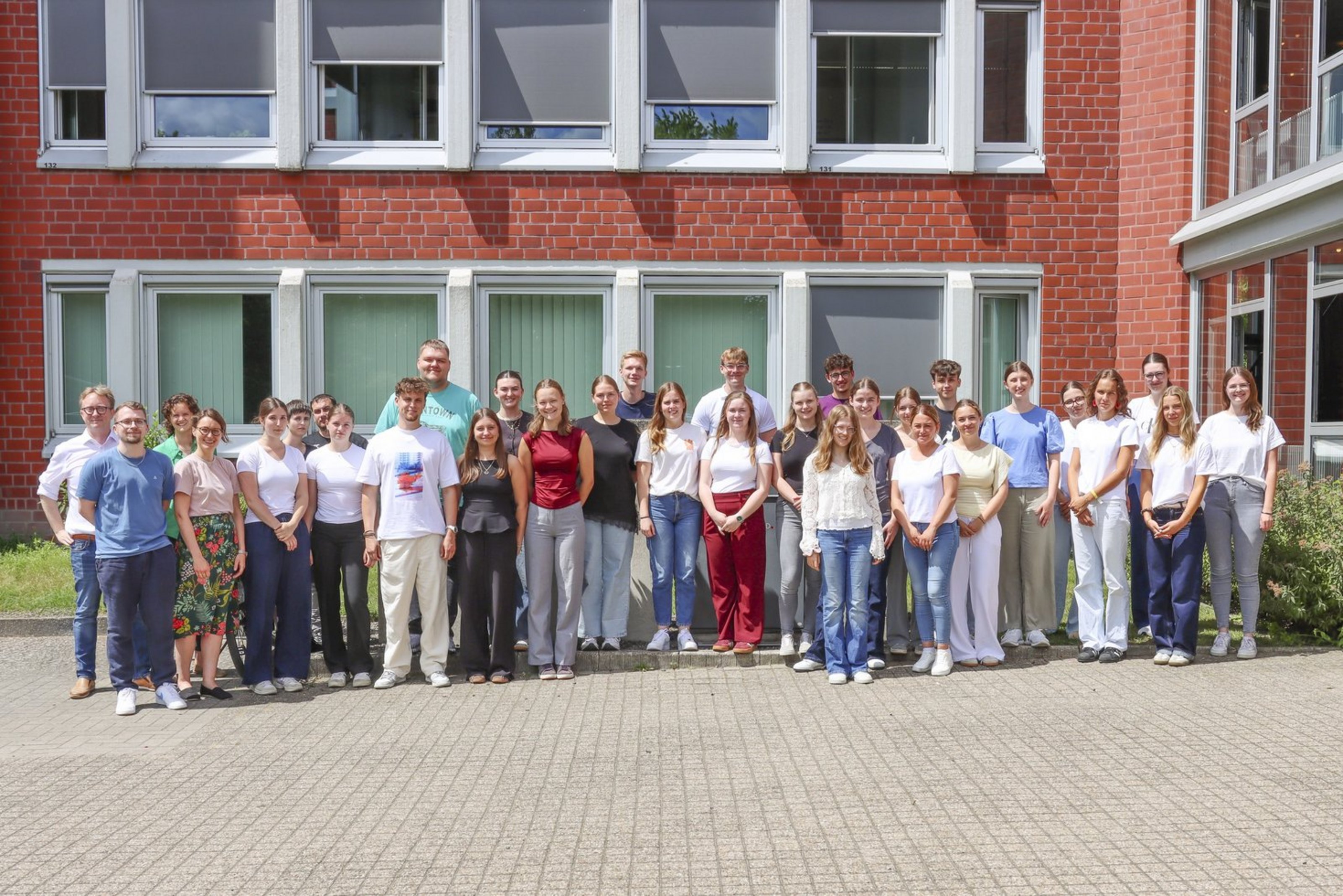 Zweiunddreißig junge Männer und Frauen stehen als Gruppe vor einem Gebäude aus rotem Backstein.