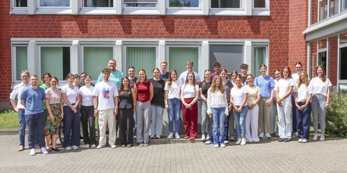 Zweiunddreißig junge Männer und Frauen stehen als Gruppe vor einem Gebäude aus rotem Backstein.