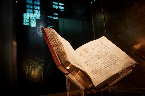 Ein altes, aufgeschlagenes Buch mit dicken, welligen Seiten liegt schräg in einer gläsernen Vitrine. Das Buch zeigt eine Seite mit lateinischem Text in feiner, brauner Schrift. 