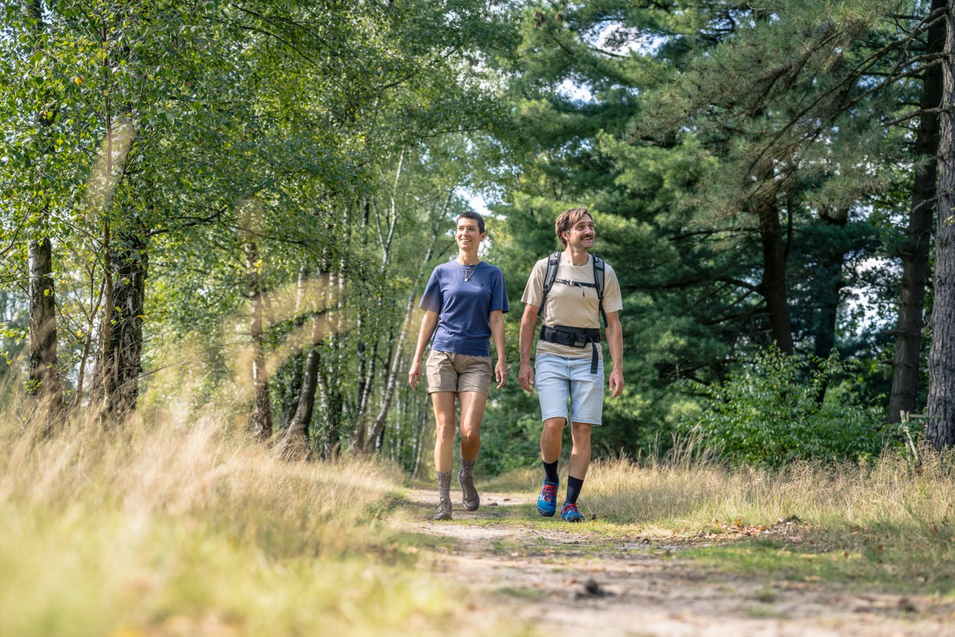 Ein Mann mit Rucksack und eine Frau wandern durch einen Wald. Sie tragen kurze Hosen und T-Shirts und schauen erfreut.