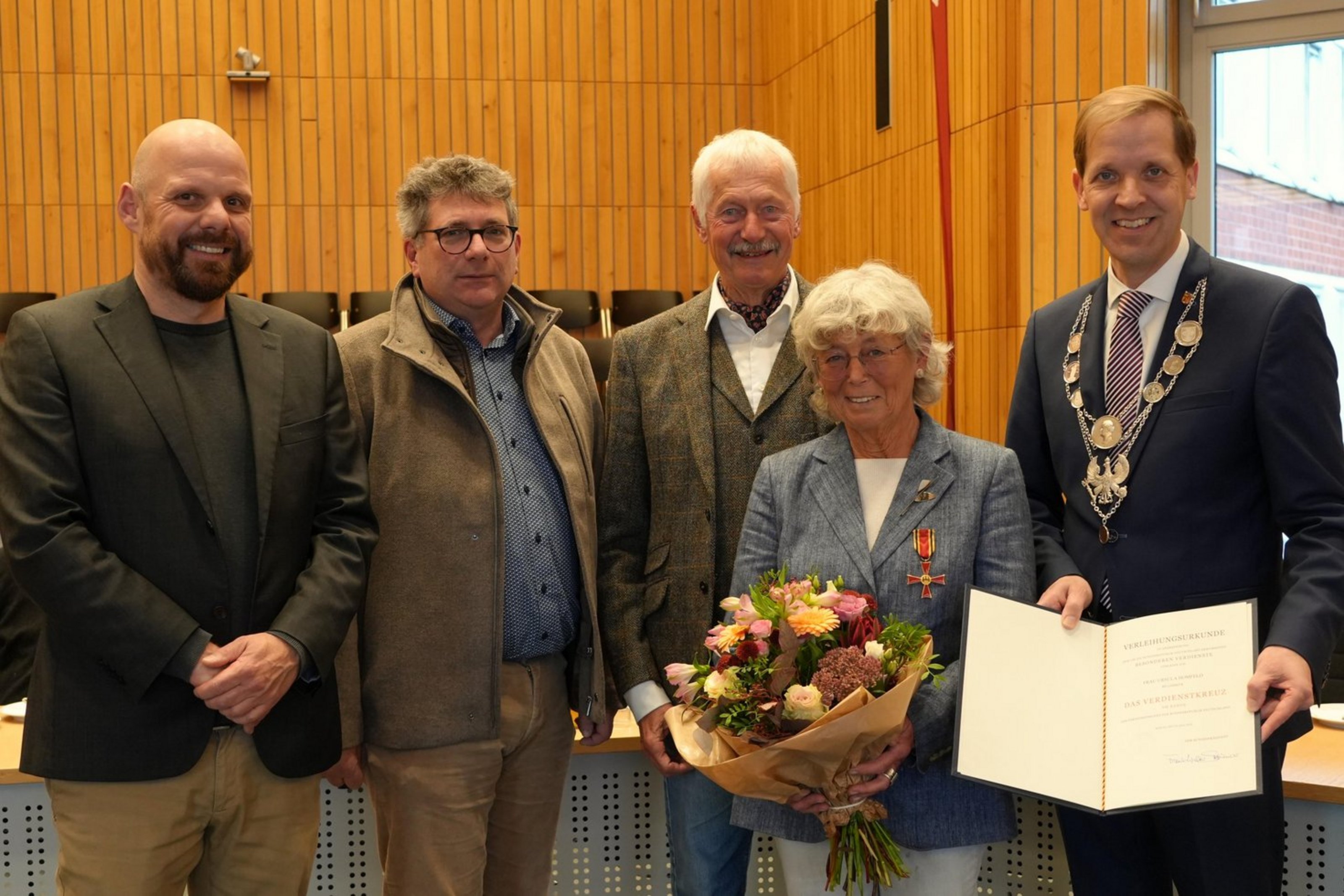 Vier Männer und eine Frau stehen in einem großen Raum mit Holzvertäfelung. Die Frau hält einen Blumenstrauß und der Mann neben ihr eine Urkundenmappe.