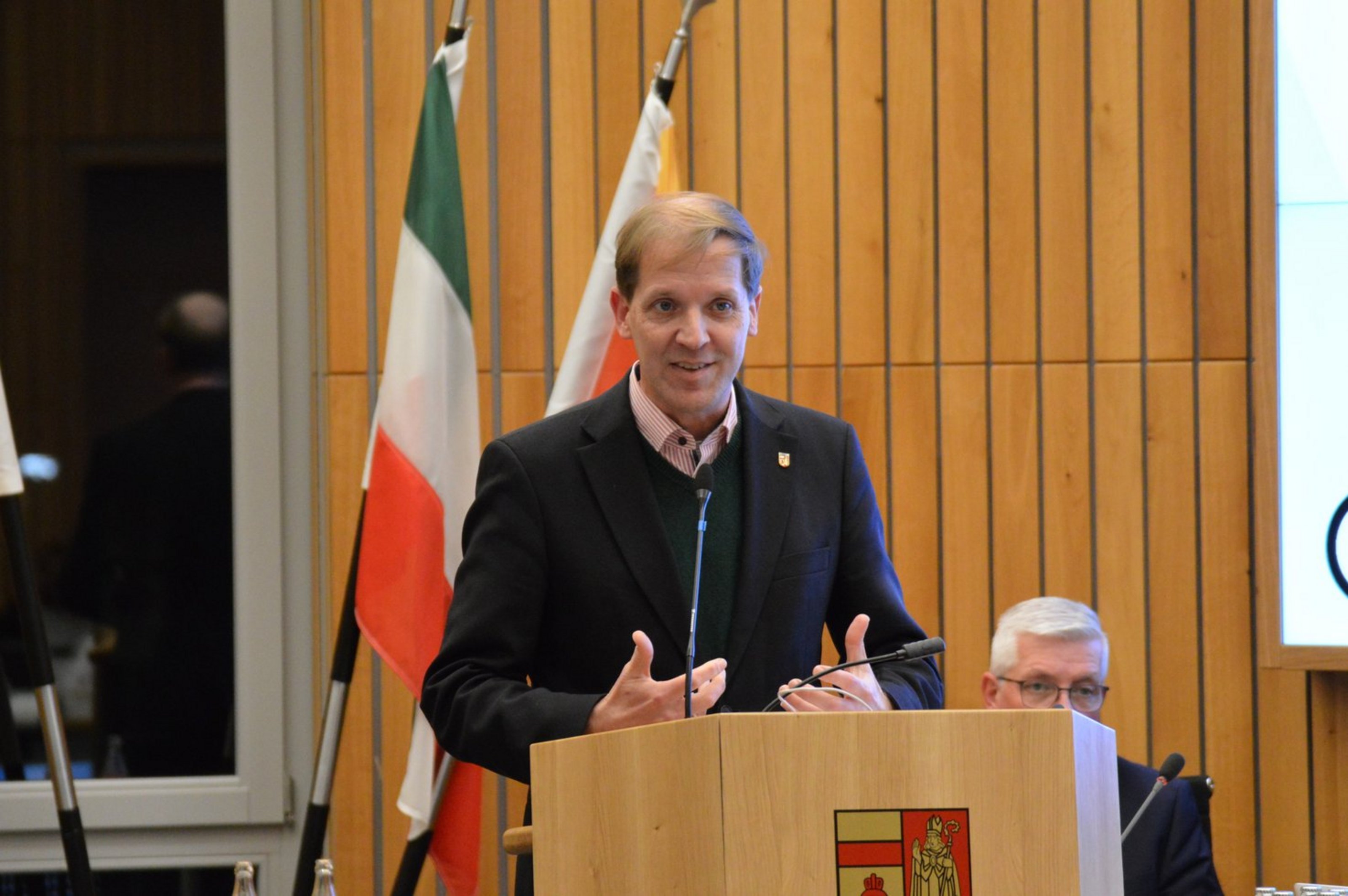 Ein Mann mit Jackett steht hinter einem REdnerpult und gestikuliert mit den Händen. Hinter ihm stehen zwei Fahnen.