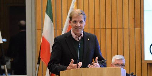 Ein Mann mit Jackett steht hinter einem REdnerpult und gestikuliert mit den Händen. Hinter ihm stehen zwei Fahnen.