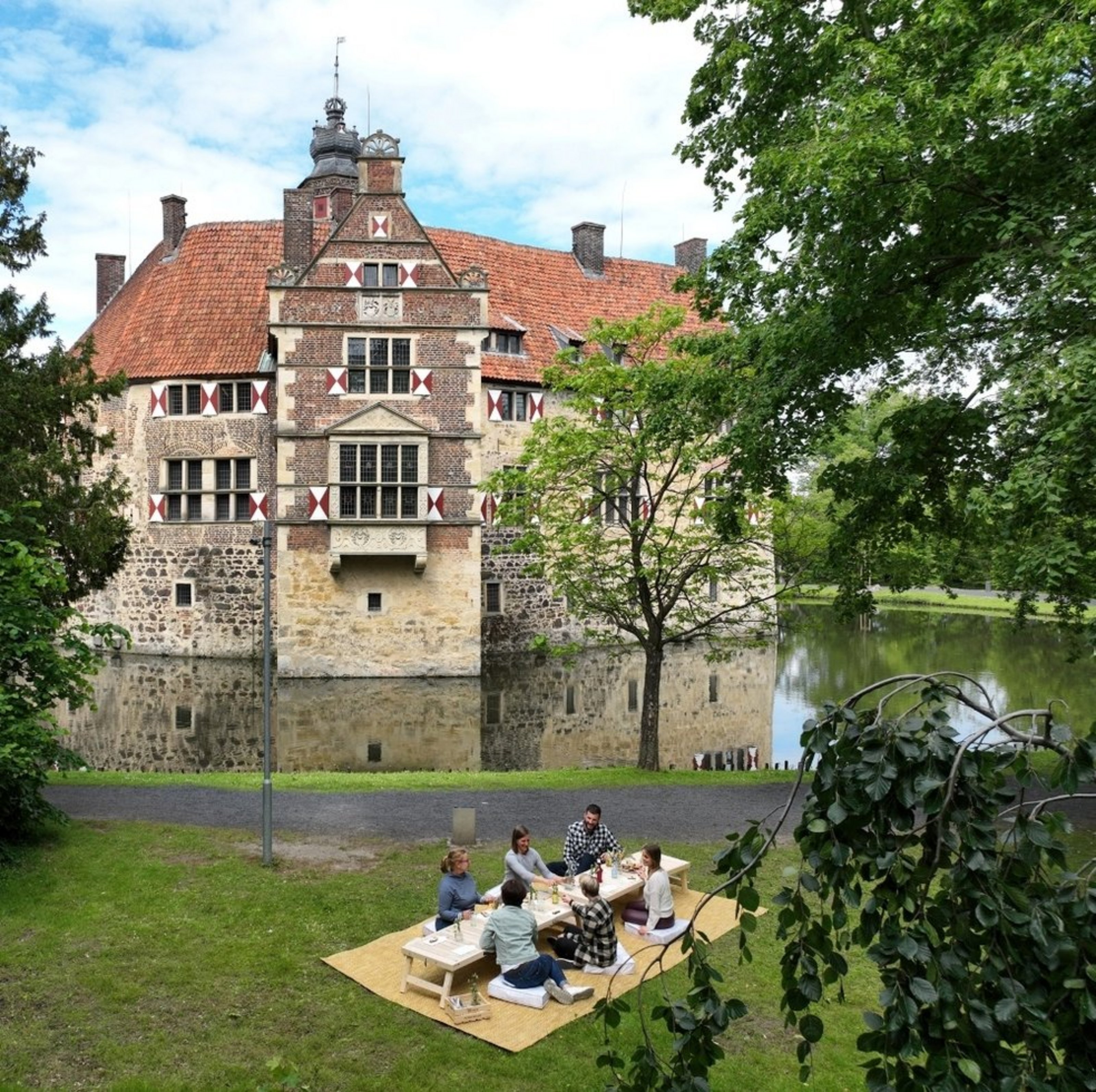 Sechs Personen sitzen an einem Picknick-Tisch auf einer grünen Wiese. Im Hintergrund ist eine Wasserburg zu sehen.