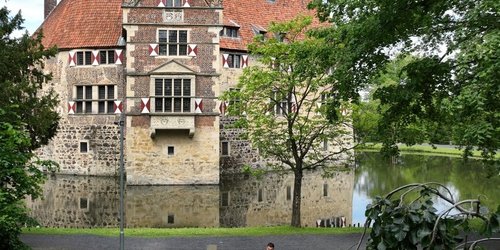 Sechs Personen sitzen an einem Picknick-Tisch auf einer grünen Wiese. Im Hintergrund ist eine Wasserburg zu sehen.