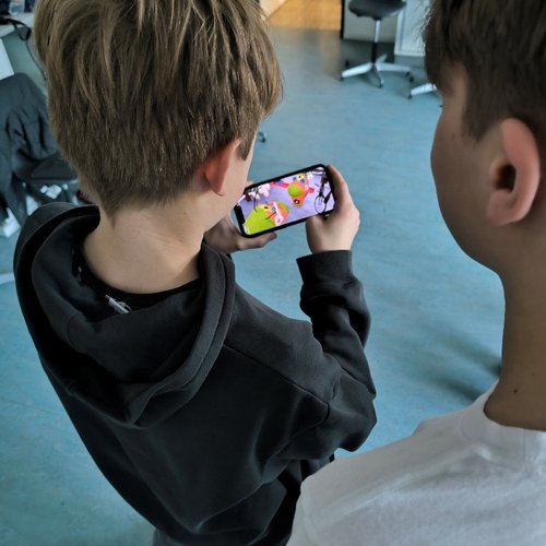 Zwei Jungen schauen auf einen Handydisplay, der zwei bunte Figuren zeigt.