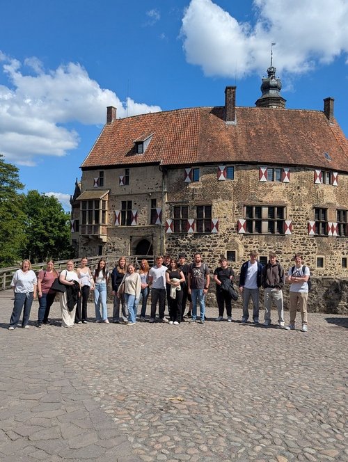 17 Personen stehen bei sonnigem Wetter im Innenhof der Vorburg von burg Vischering. Im Hintergrund sind blauer Himmel und die Burg Vischering zu sehen.
