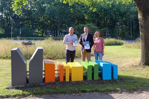 Auf einer Blumenwiese hinter einer bunten, Kniehohen Skulptur stehen zwei Männer und eine Frau. Die beiden Männer halten eine Broschüre in den Händen.