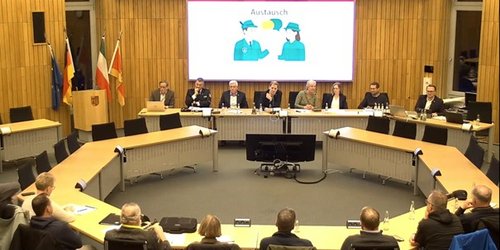 Im Sitzungssaal sitzen 16 Personen im Kreis an Tischen. Auf der