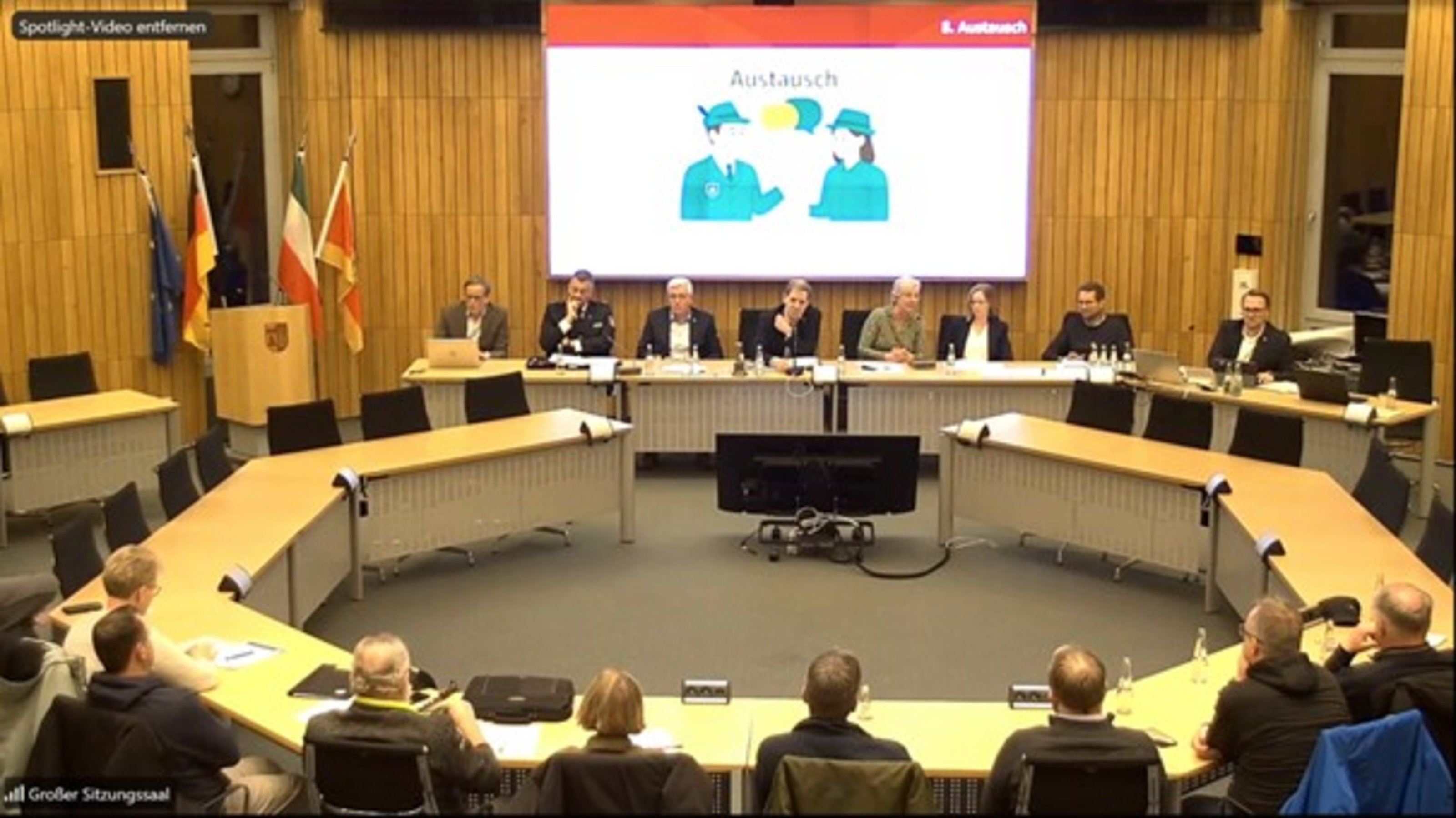 Im Sitzungssaal sitzen 16 Personen im Kreis an Tischen. Auf der