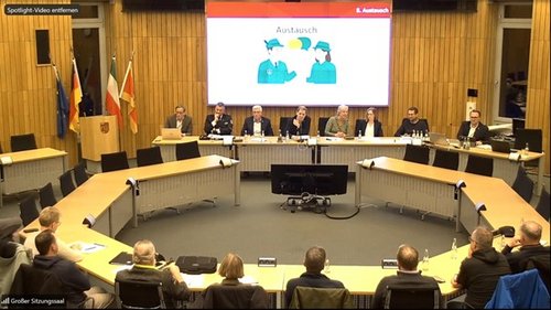 Im Sitzungssaal sitzen 16 Personen im Kreis an Tischen. Auf der