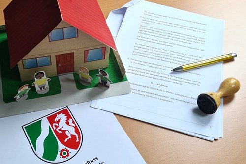 Ein Schreibtisch mit Papierdokumenten mit Landeswappen auf denen ein Modellhaus steht und ein Kugelschreiber sowie ein Stempel liegen.