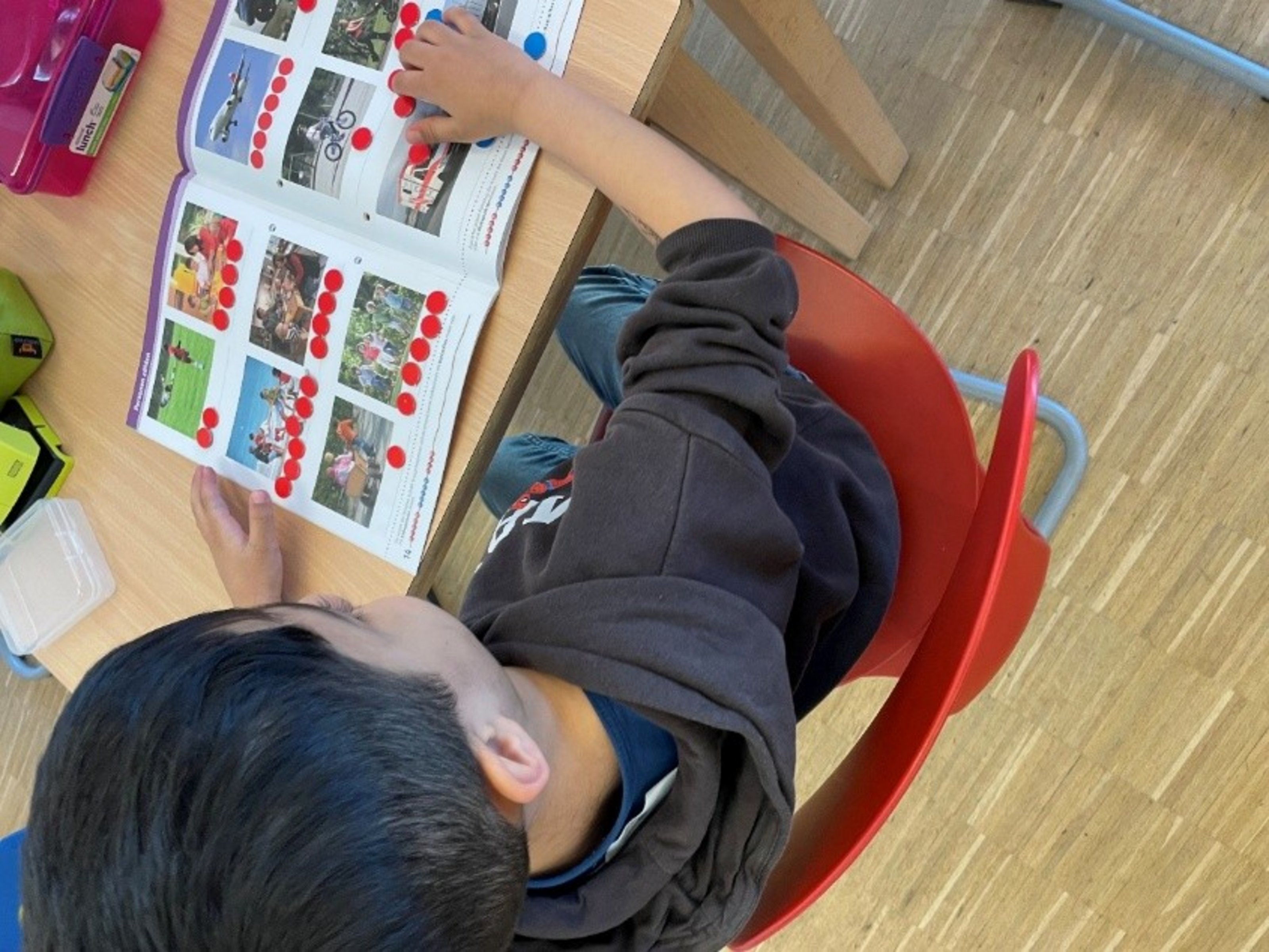 Ein Junge sitzt auf einem Stuhl an einem Tisch und schaut sich ein Buch an, das aufgeschlagen auf dem Tisch liegt.