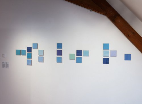 An einer weißen Wand hängen 20 Quadrate in unterschiedlichen Blautönen.