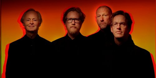 Vier Männer in schwarzen Oberteilen stehen vor einem rot-gelbem Hintergrund. Die Konturen der Männer sind verschwommen.