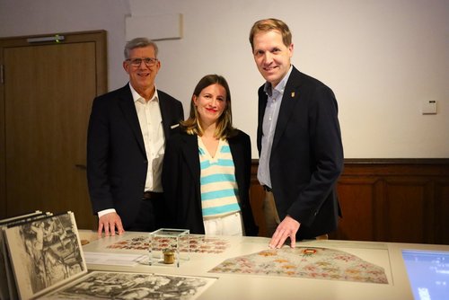 Drei Personen stehen in der Ausstellung der Burg Vischering und präsentieren historische Exponate, darunter ein Petschaftsiegel, das auf einem Tisch vor ihnen liegt.