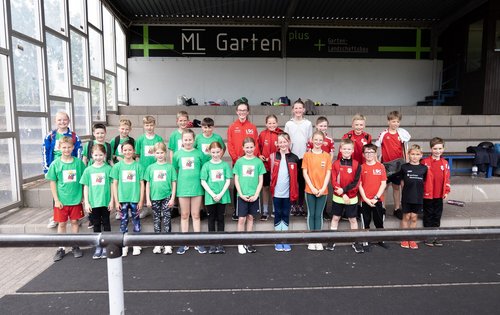 Eine Gruppe von 25 Kindern steht in zwei reihen vor einer Tribüne . Die Kinder tragen Sport-Klamotten.