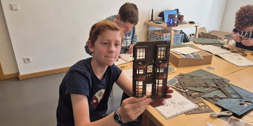 Ein Junge sitzt mit zwei anderen Jungen an einem Gruppentisch, auf dem Holzteile liegen. Der eine Junge hält eine Miniaturhausfront in die Kamera.