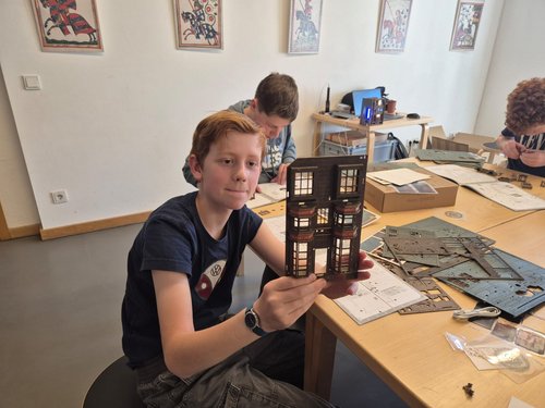 Ein Junge sitzt mit zwei anderen Jungen an einem Gruppentisch, auf dem Holzteile liegen. Der eine Junge hält eine Miniaturhausfront in die Kamera.