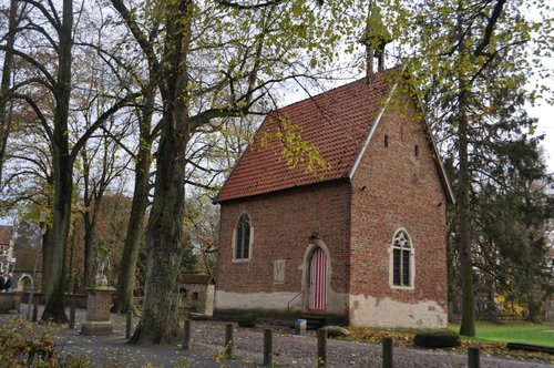 Eine kleine Backsteinkapelle mit rotem Satteldach steht inmitten hoher, laubtragender Bäume. Die Kapelle hat spitzbogige Fenster mit weißem Rahmen und eine rot-weiß gestreifte Holztür an der Frontseite. Davor führen wenige Stufen zum Eingang. Links im Bild ist eine Statue auf einem gemauerten Sockel zu sehen. Die Umgebung wirkt herbstlich, mit gelb verfärbtem Laub an den Bäumen und am Boden.