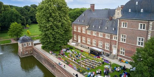 Im Schlossgarten vor einem Schloss stehen auf einer Rasenfläche eine Bühne und viele Liegestühle, die zur Bühne ausgerichtet sind. Auch an umliegenden Picknickbänken sitzen Menschen. Der Garten ist von einem Wassergraben umgeben.