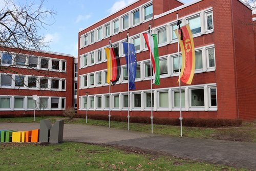 Vor einem roten Verwaltungsgebäude mit vielen weißen Fenstern sind vier Flaggenmasten aufgestellt. An den Masten wehen (von links nach rechts) die deutsche Flagge auf Halbmast, die Flagge der Europäischen Union, die Flagge des Bundeslands Nordrhein-Westfalen, eine gelb-rote Flagge mit einem Wappen – alle ebenfalls auf Halbmast. Links im Vordergrund steht eine bunte Skulptur mit Buchstaben. Der Himmel ist leicht bewölkt, es ist Tag.