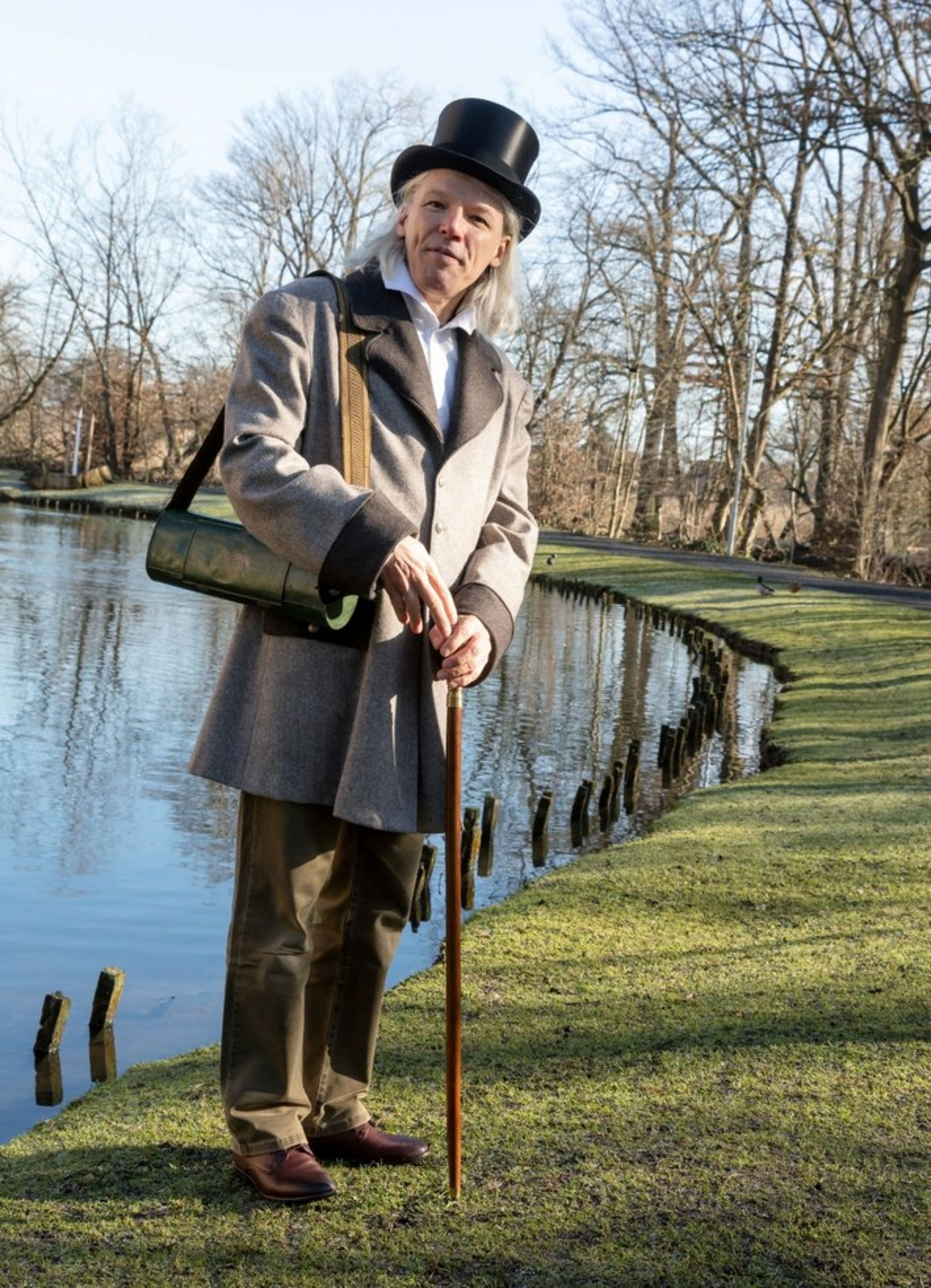 Ein Mann in historischer Kleidung steht mit Krückstock an einem See. Er trägt einen Zylinder und einen Anorak. 