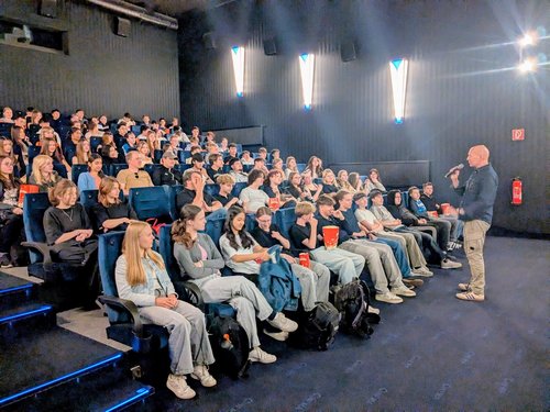 Ein vollbesetzter Kinosaal mit Blick auf die Sitzreihen. Junge Menschen sitzen in den Kinosesseln und haben vereinzelnt rote Popcorntüten auf dem Schoß. Vor den Sitzreihen steht ein Mann mit Mikrofon, der ins Publikum blickt. Der Boden ist blau, die Wände auch.