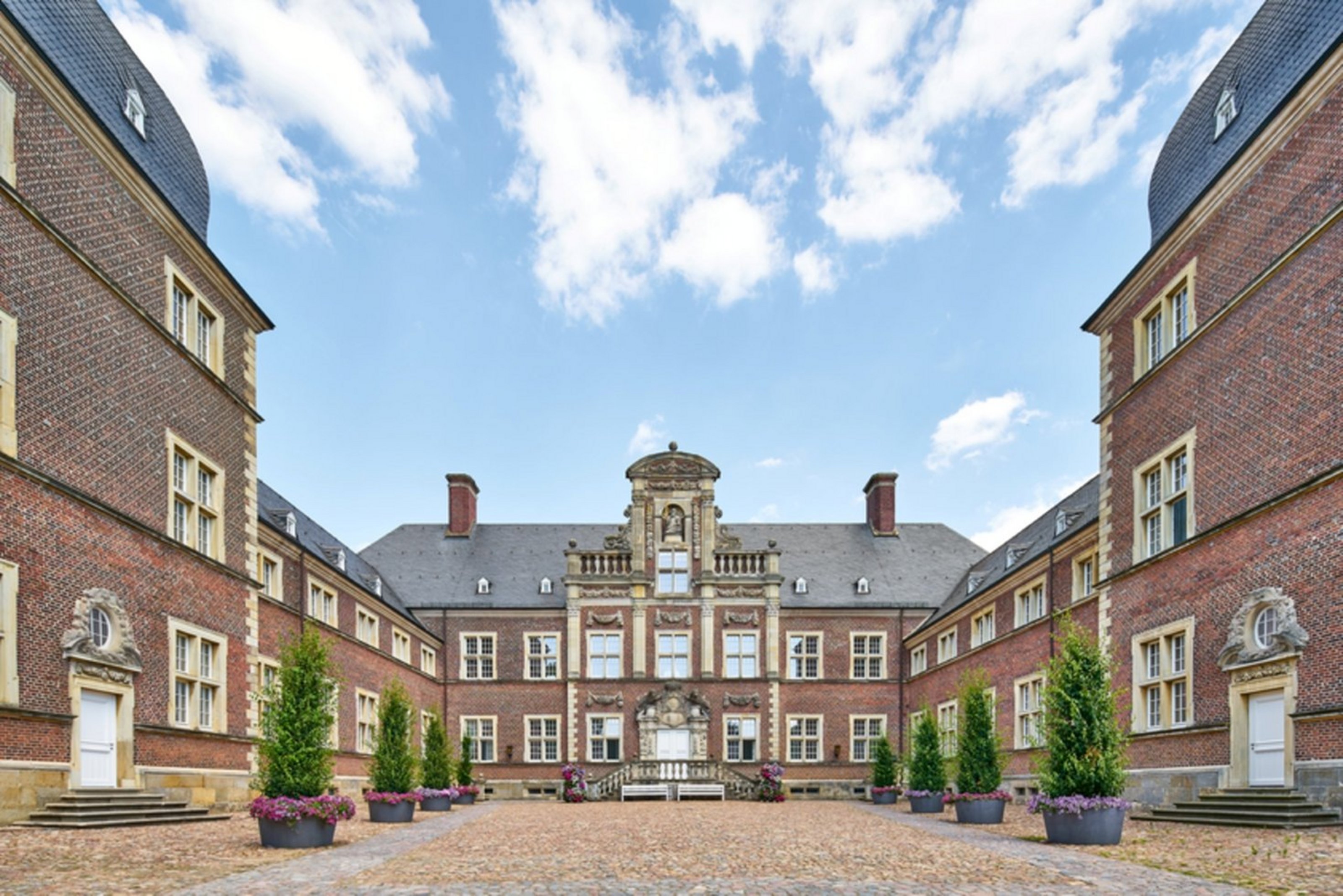 Der Innenhof des Schlosses Ahaus (Bildquelle: Münsterland e.V., Philipp Fölting) Innenhof eines historischen Schlosses mit symmetrischer Backsteinarchitektur. Drei Gebäudeflügel rahmen den gepflasterten Hof ein, in dessen Mitte Pflanzen in großen Kübeln stehen. Die Fassade des mittleren Gebäudeflügels ist besonders aufwendig verziert, mit einem zentralen Turmerker, klassizistischen Statuen und Balustraden. Der Himmel ist blau mit einigen weißen Wolken