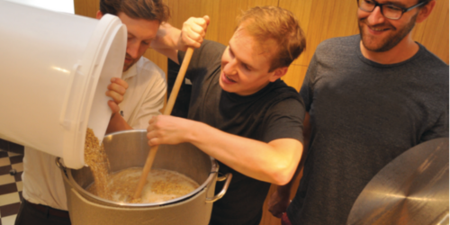 Drei Männer stehen an einem großen Topf. Einer kippt Getreide aus einem Eiimer in den Topf, einer rührt mit einem Holzlöffel und einer schaut zu.
