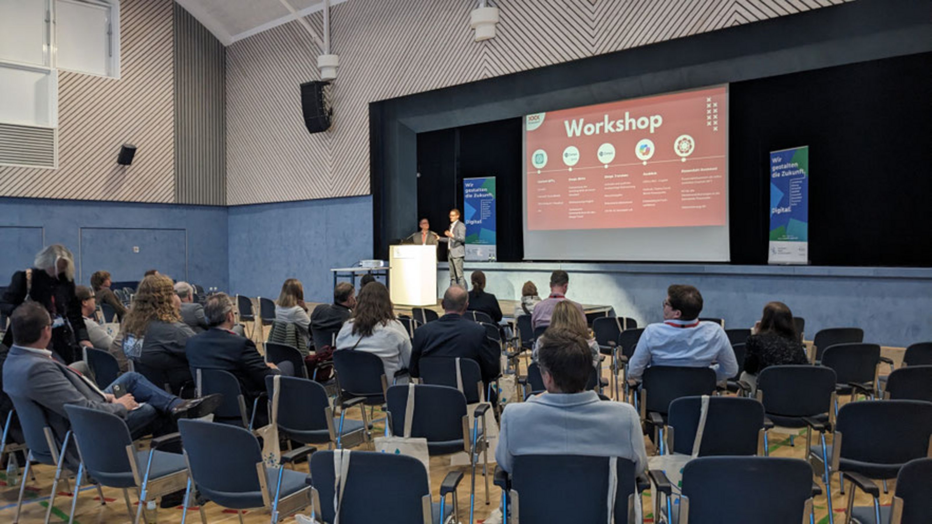 Blick in einen Veranstaltungssaal mit sitzenden Teilnehmenden. Auf der Bühne präsentieren zwei Personen einen Workshop, sichtbar ist eine Folie mit der Überschrift ‚Workshop‘.