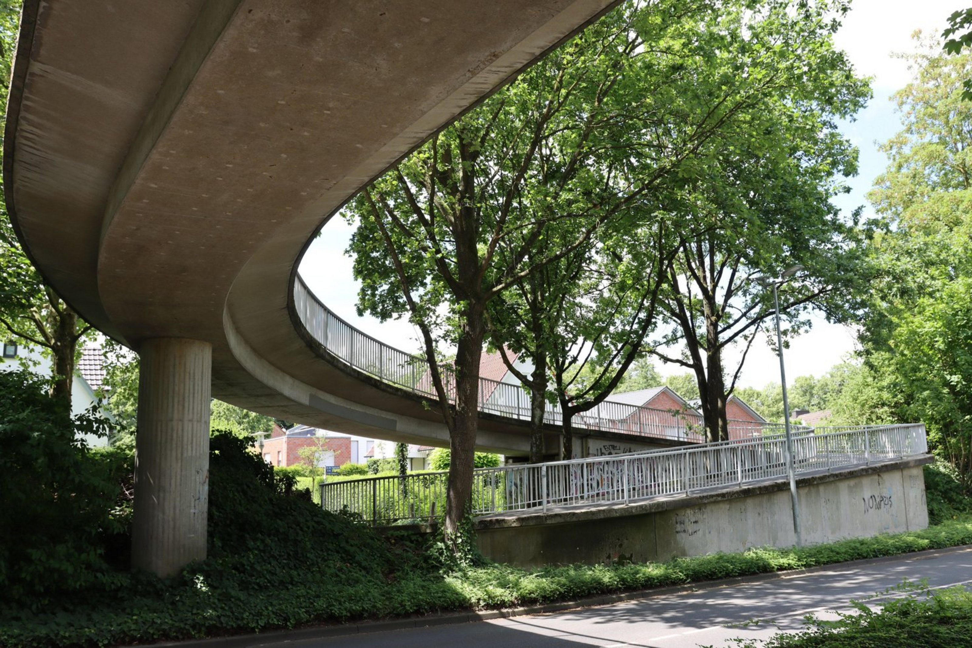 Eine Betonbrück überquert in einem geschwungenen Bogen eine Straße. Im Hintergrund stehen Bäume.