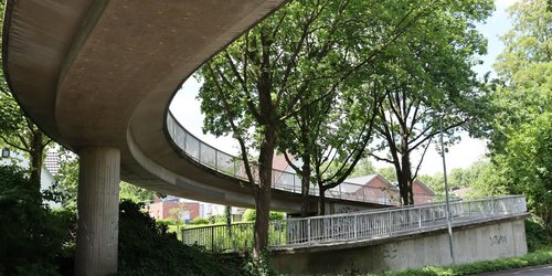 Eine Betonbrück überquert in einem geschwungenen Bogen eine Straße. Im Hintergrund stehen Bäume.