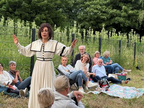 Auf einer Wiese vor einem Zaun steht eine Frau in einem langen, weißen Kleid mit schwarzen Akzenten und spricht mit ausgebreiteten Armen. Im Hintergrund sitzen mehrere Menschen unterschiedlichen Alters entspannt auf Picknickdecken und Klappstühlen, einige hören aufmerksam zu, andere schauen gedankenverloren. Hinter dem Zaun wachsen hohe, blühende Pflanzen, dahinter dichter grüner Wald.