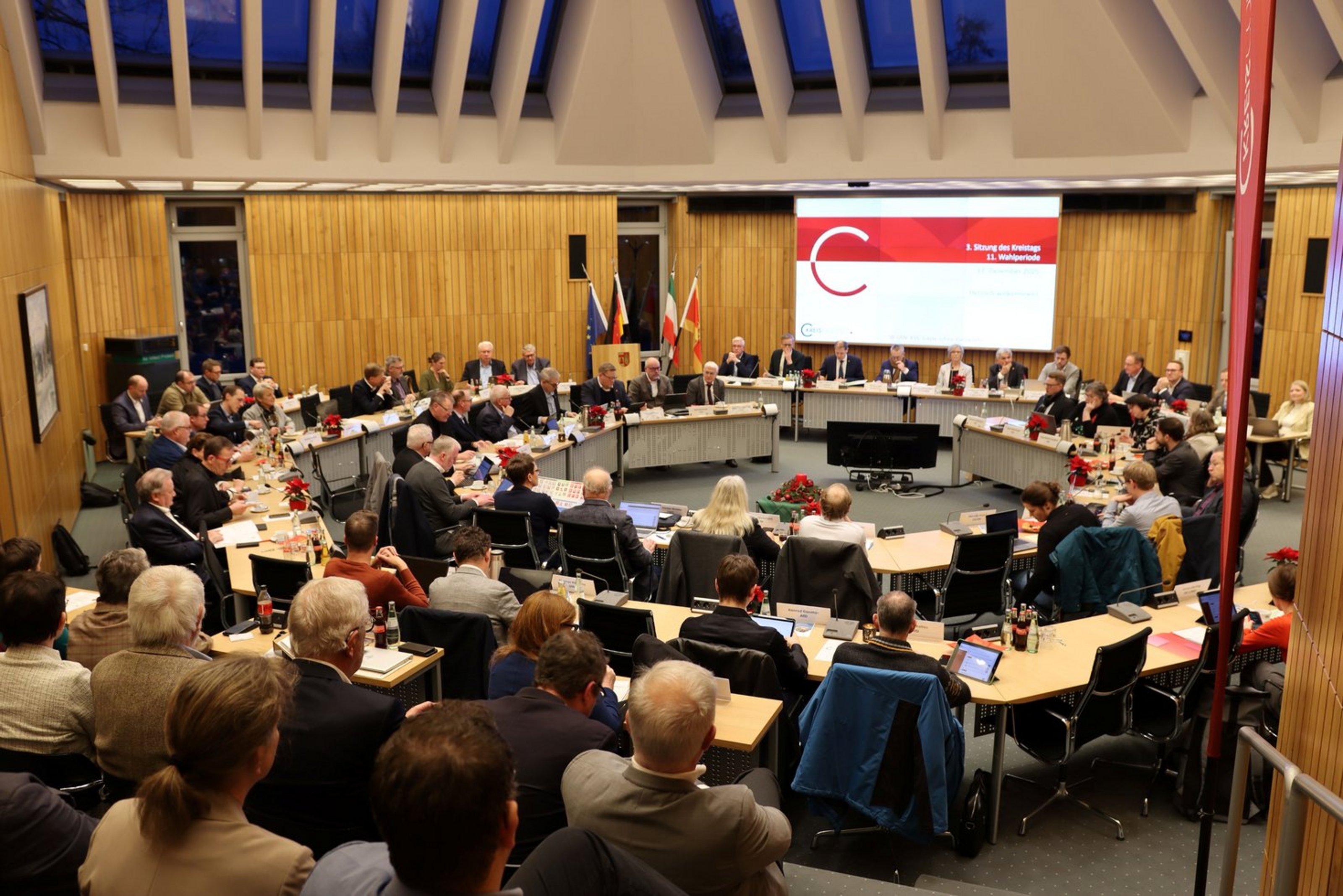 In einem Sitzungssaal befinden sich zwei Tischkreise und eine Tribüne an und auf der Personen sitzen und Richtung eines großen Bildschirmes schauen.