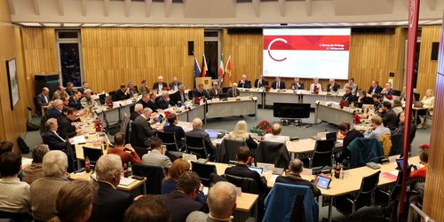 In einem Sitzungssaal befinden sich zwei Tischkreise und eine Tribüne an und auf der Personen sitzen und Richtung eines großen Bildschirmes schauen.