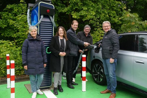 Fünf Personen stehen an einer e-Ladesäule neben einem Auto. Drei Personen betanken das Auto.