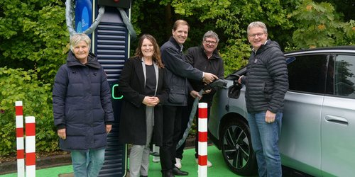 Fünf Personen stehen an einer e-Ladesäule neben einem Auto. Drei Personen betanken das Auto.