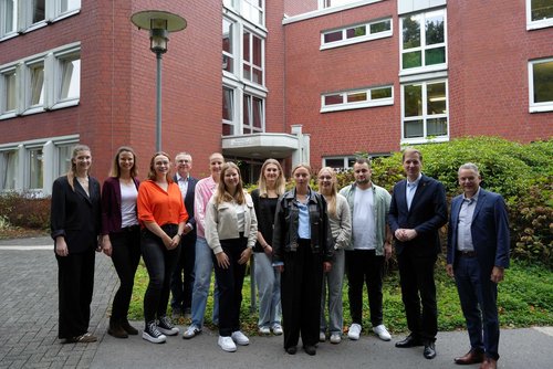 Eine Gruppe Männer und Frauen unterschiedlichen Alters steht vor einem Gebüsch. Im Hintergrund ist ein rotes Backsteingebäude.