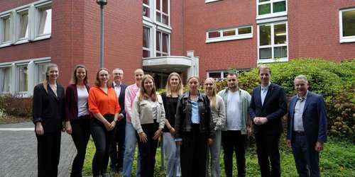 Eine Gruppe Männer und Frauen unterschiedlichen Alters steht vor einem Gebüsch. Im Hintergrund ist ein rotes Backsteingebäude.