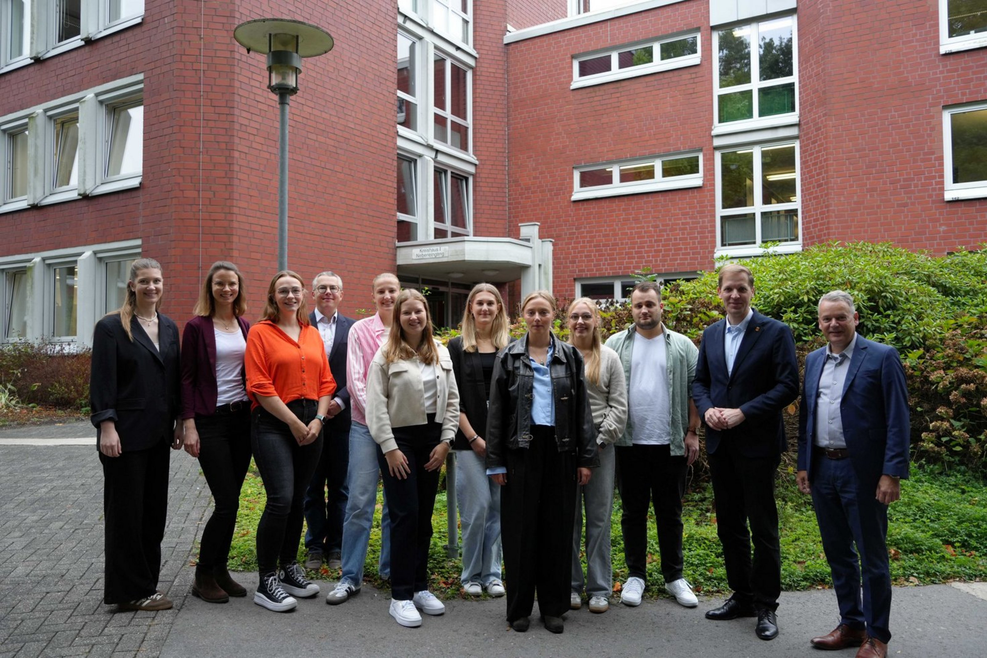 Eine Gruppe Männer und Frauen unterschiedlichen Alters steht vor einem Gebüsch. Im Hintergrund ist ein rotes Backsteingebäude.