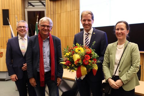 Vier Personen stehen im Sitzungssaal des Kreishauses. Kreiswahlleiter gratuliert dem wiedergewählten Landrat, der einen großen Blumenstrauß hält. Neben ihm stehen sein Gegenkandidat und seine Frau.