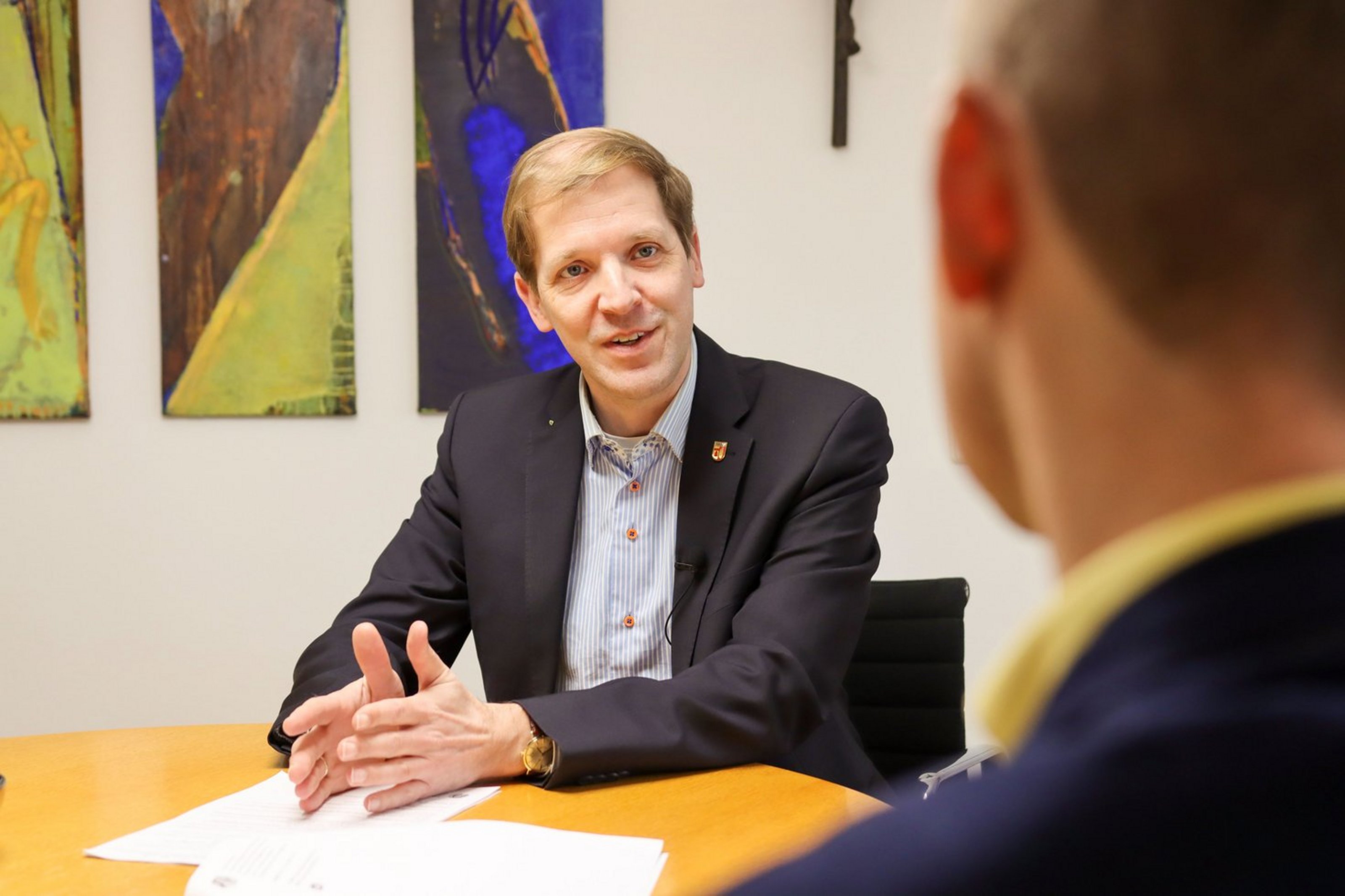 Ein Mann im Anzug sitzt an einem Besprechungstisch und spricht mit einer Person, die im Vordergrund von hinten zu sehen ist. Er gestikuliert mit den Händen und blickt sein Gegenüber freundlich an. Im Hintergrund hängen farbige Bilder an der Wand.