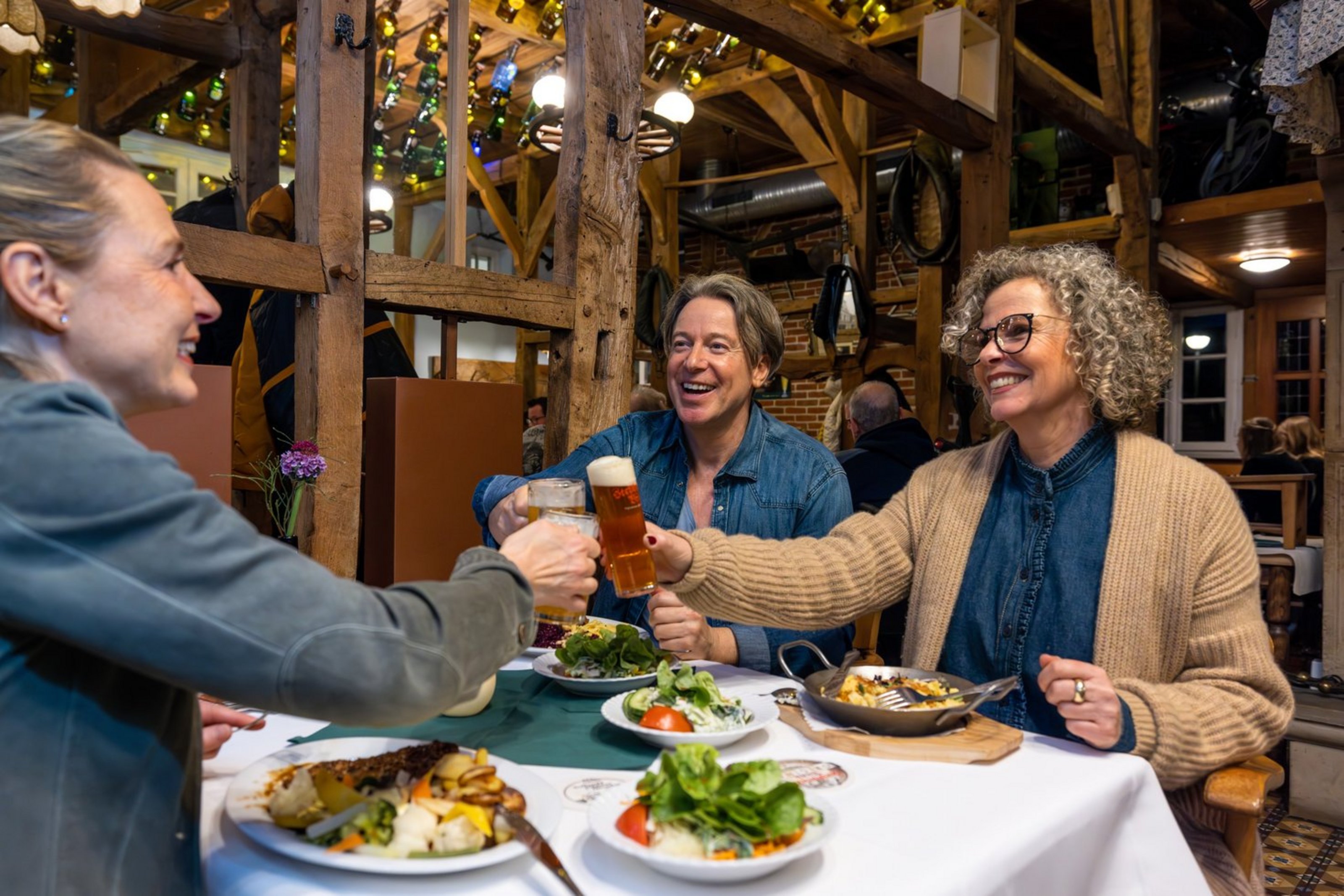 Drei Personen sitzen an einem Tisch in einem Lokal und stoßen mit Biergläsern an.
